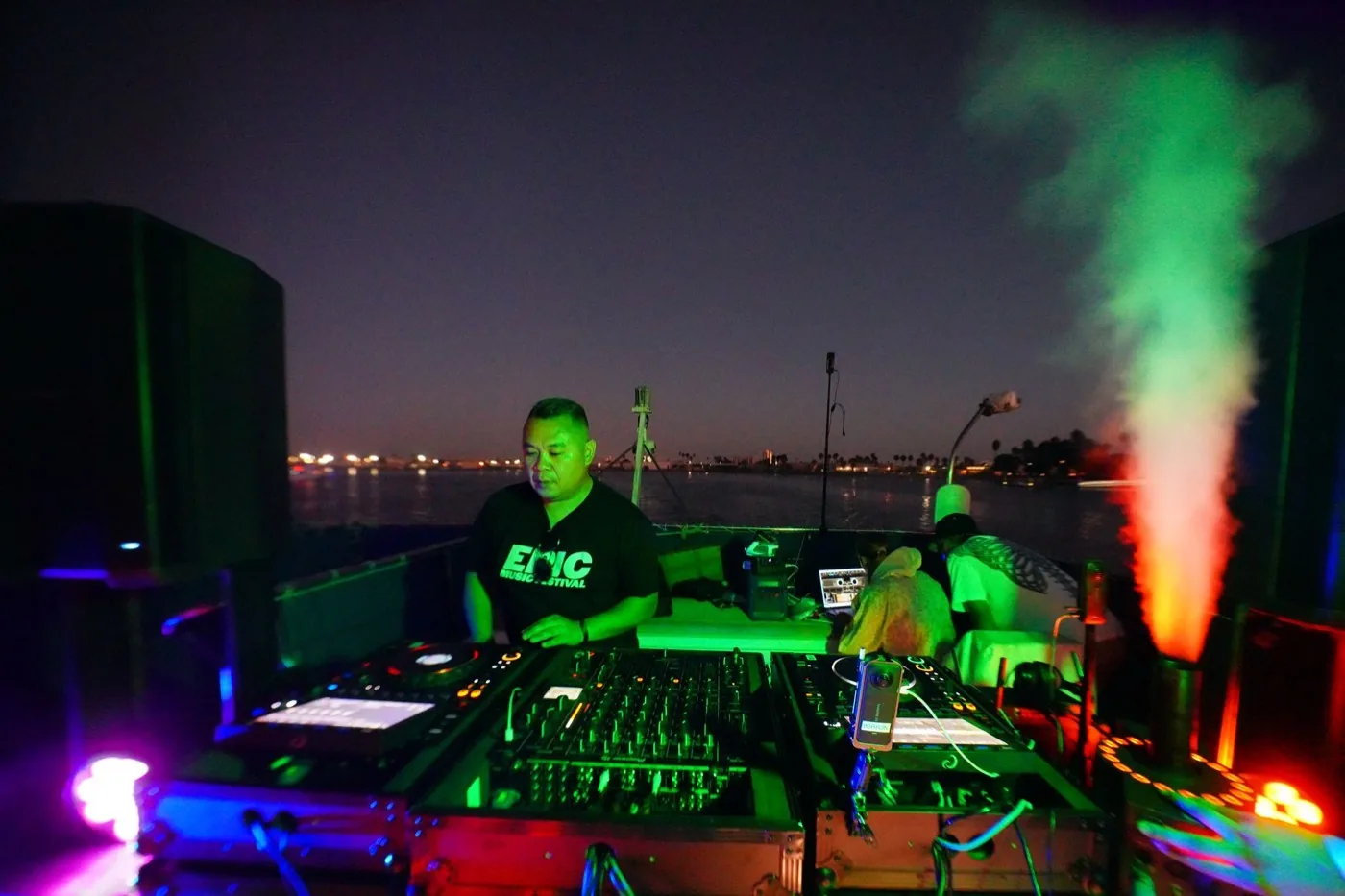 DJ in an EPIC Music Festival shirt playing on the boat deck at night, CO2 firing beside the booth and shore lights across the water