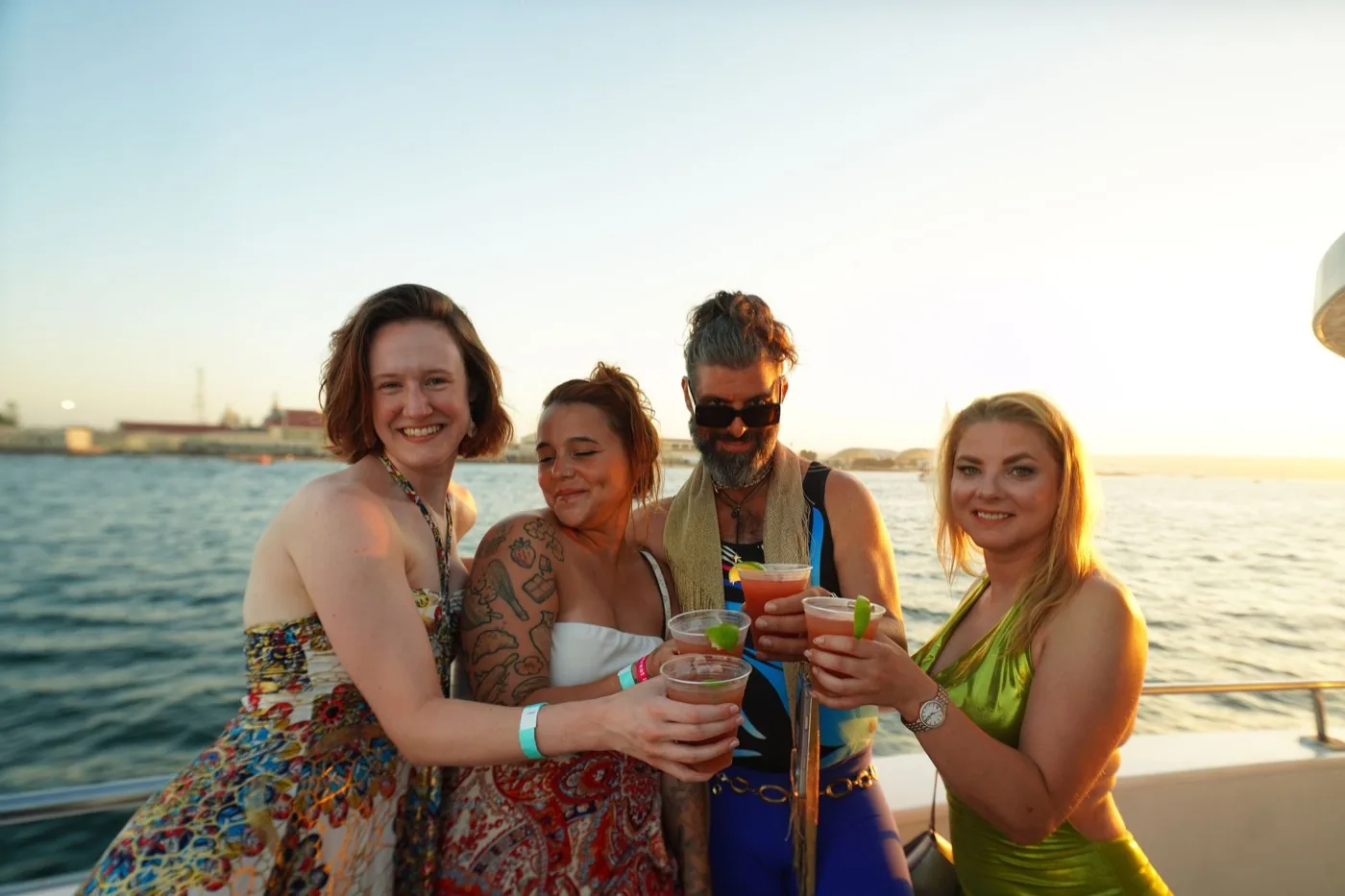 Four people on the top deck of a boat at sunset holding drinks, wearing event wristbands