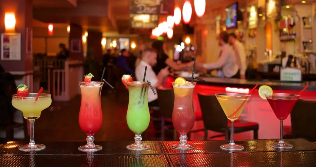 A row of cocktails on a lit bar with staff working behind it